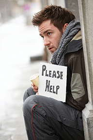 Man sitting outdoors with a Please Help sign, wearing a scarf and holding a cup, appearing in need of assistance.