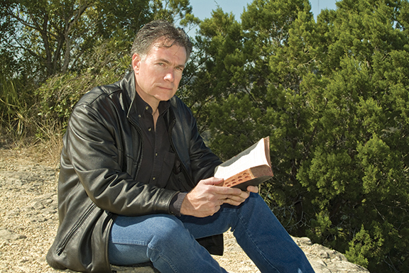 Man in a leather jacket reading a book outdoors near greenery, sitting on a rocky surface.