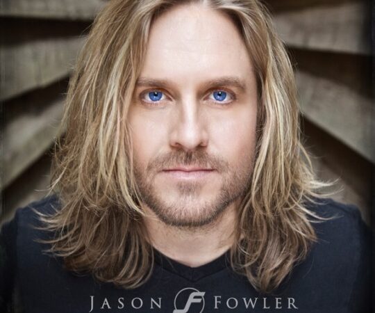 Portrait of a person with long blonde hair and blue eyes, set against a neutral background.