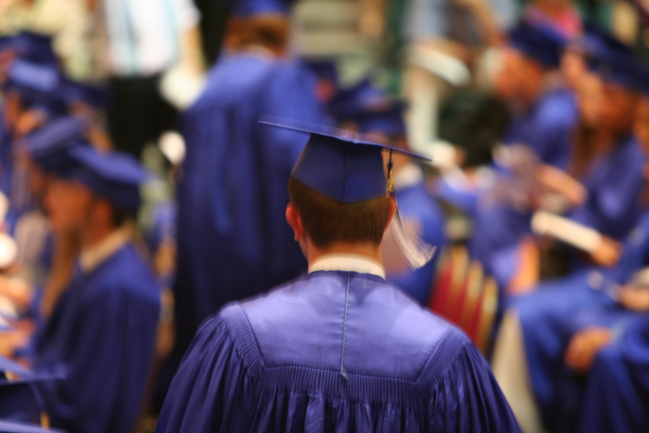 Graduate in blue cap and gown at commencement ceremony, facing stage.