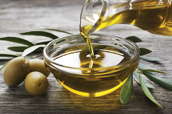 Pouring olive oil into a bowl with fresh olives and leaves on a wooden table.