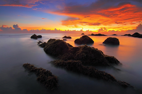 Sunset over rocky shoreline with vibrant orange and purple sky, reflecting on calm sea water.