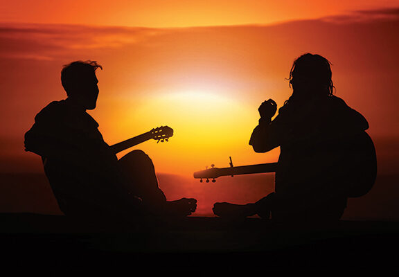 Silhouetted musicians playing guitars at sunset, creating a serene, musical atmosphere against a vibrant orange sky.