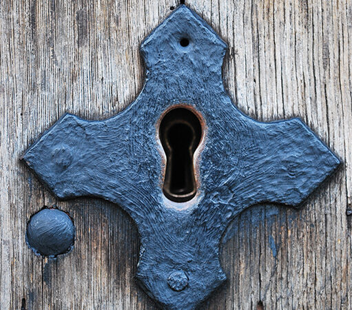Ornate vintage keyhole in wood door, shaped like a cross, with weathered blue metal detail.