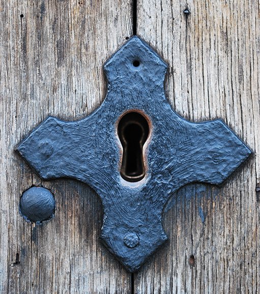 Ornate vintage keyhole in wood door, shaped like a cross, with weathered blue metal detail.