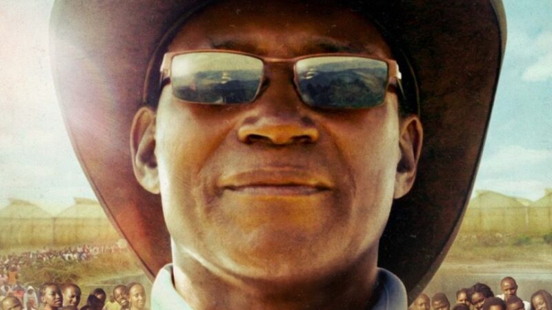 Man in sunglasses and hat with crowds in the background under a sunny sky.