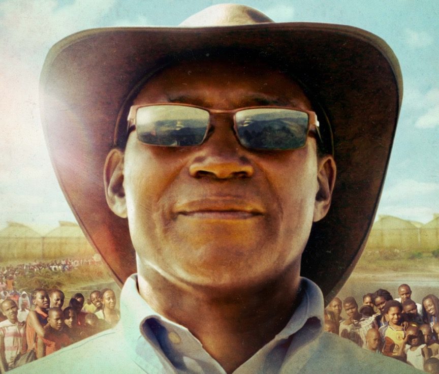 Man in sunglasses and hat with crowds in the background under a sunny sky.