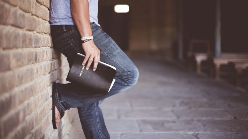 Person leaning on wall holding a Bible, wearing jeans and flip-flops, suggesting reflection or spirituality.
