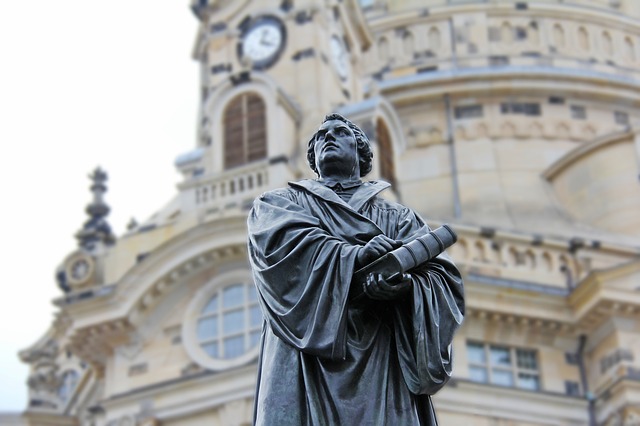 Statue of historical figure with book in front of ornate architecture with clock tower in background.