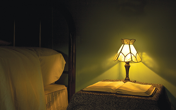 Bedside table with a warm-lit lamp illuminating an open book in a cozy bedroom setting.
