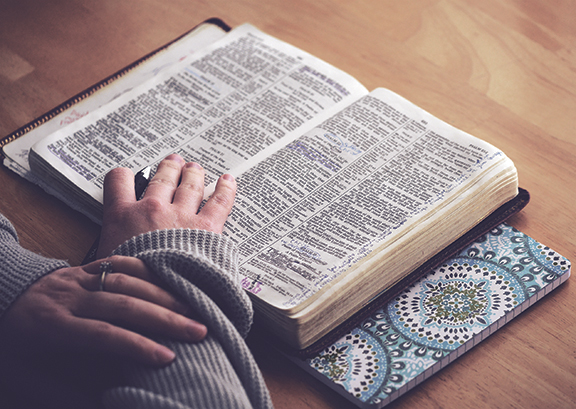 Person reading an open Bible on a table with a patterned bookmark visible.