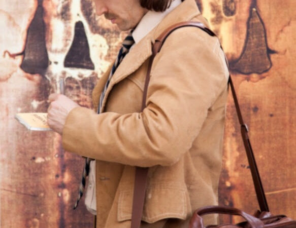 Person reading notes, carrying a leather bag, in front of a vintage, patterned background.