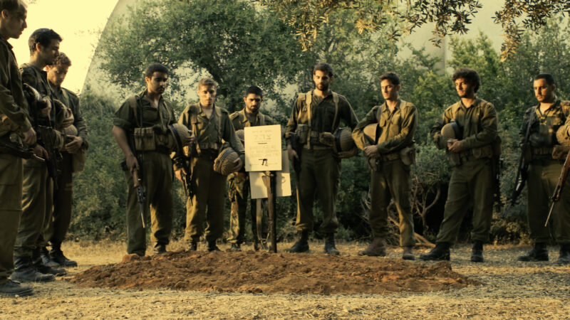 Soldiers in uniform stand solemnly in a field, paying respects at a grave.