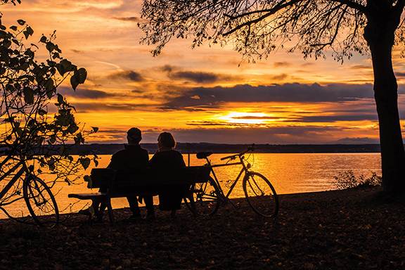 Two people sit on a bench by the lake, watching a vibrant sunset with bicycles nearby.