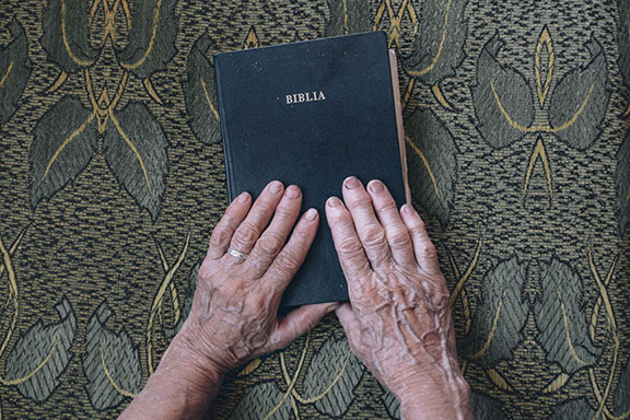 Elderly hands holding a Bible on a patterned fabric background, symbolizing faith and spirituality.