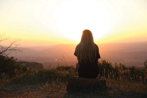Person sitting on hilltop watching sunset over landscape, serene view, nature, peaceful moment.