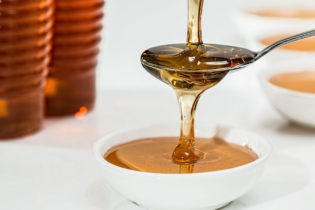 Spoon pouring golden honey into a white bowl; honey jars in the background. Perfect for natural sweetener content.