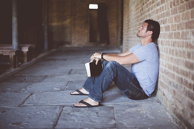 Man sitting against brick wall with a book, looking thoughtful. Casual outfit, serene atmosphere in covered walkway.