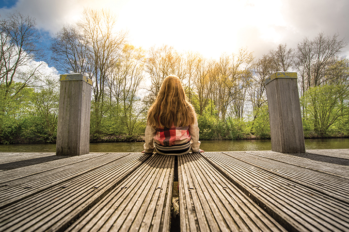 Person sitting on a wooden pier, gazing at a serene river under a bright sky, surrounded by trees in spring.