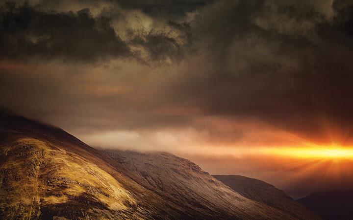 Dramatic mountain landscape at sunset with dark clouds and a vibrant orange sun peeking through.