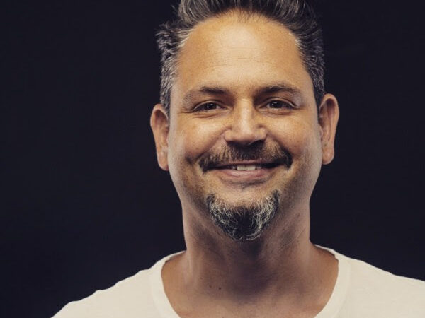 Smiling man with styled hair and goatee, wearing a white shirt against a dark background.