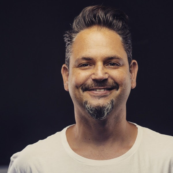 Smiling man with styled hair and goatee, wearing a white shirt against a dark background.