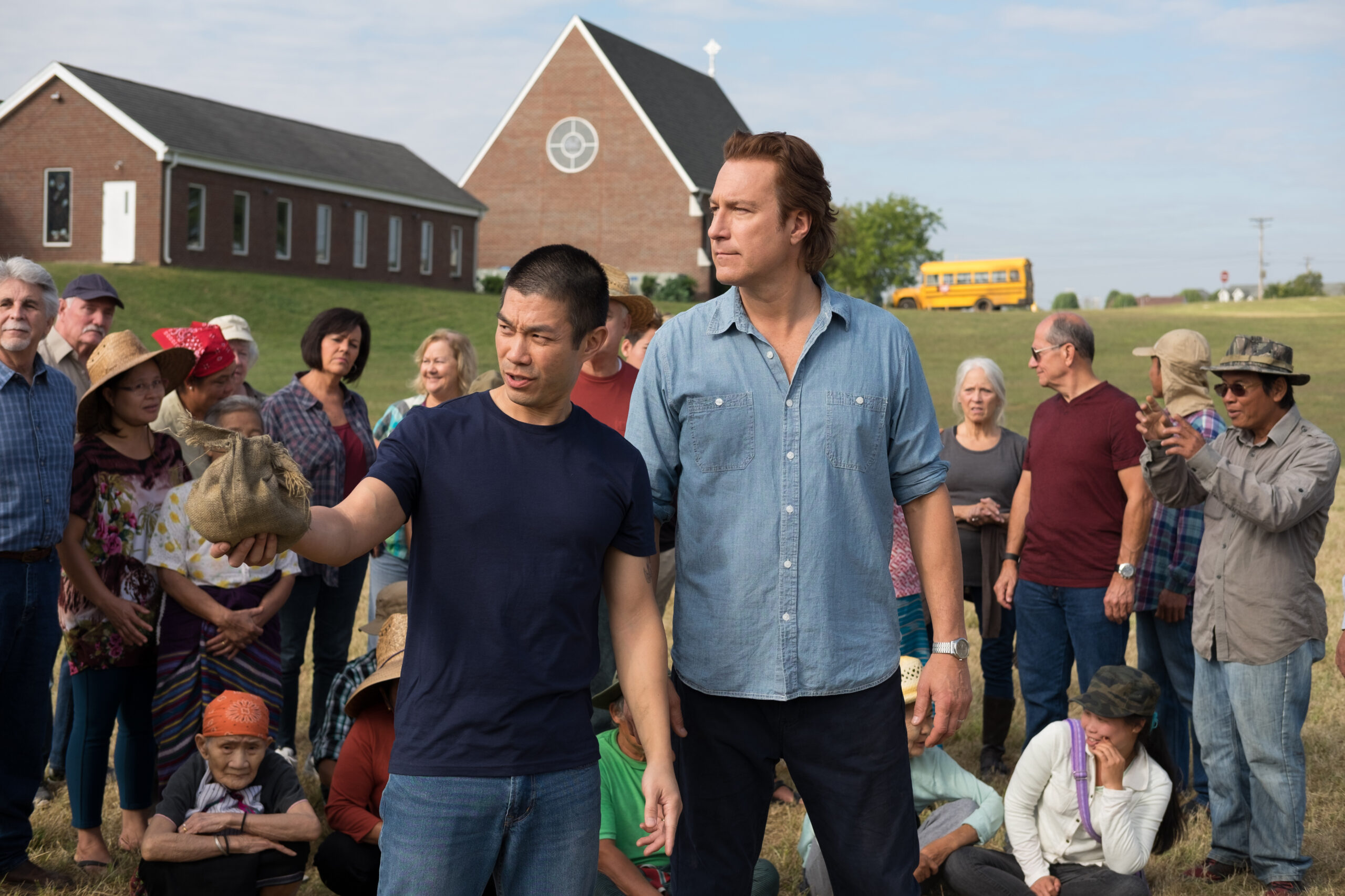Community gathering in front of a church, man holding a burlap sack, diverse group listening attentively, bus in background.