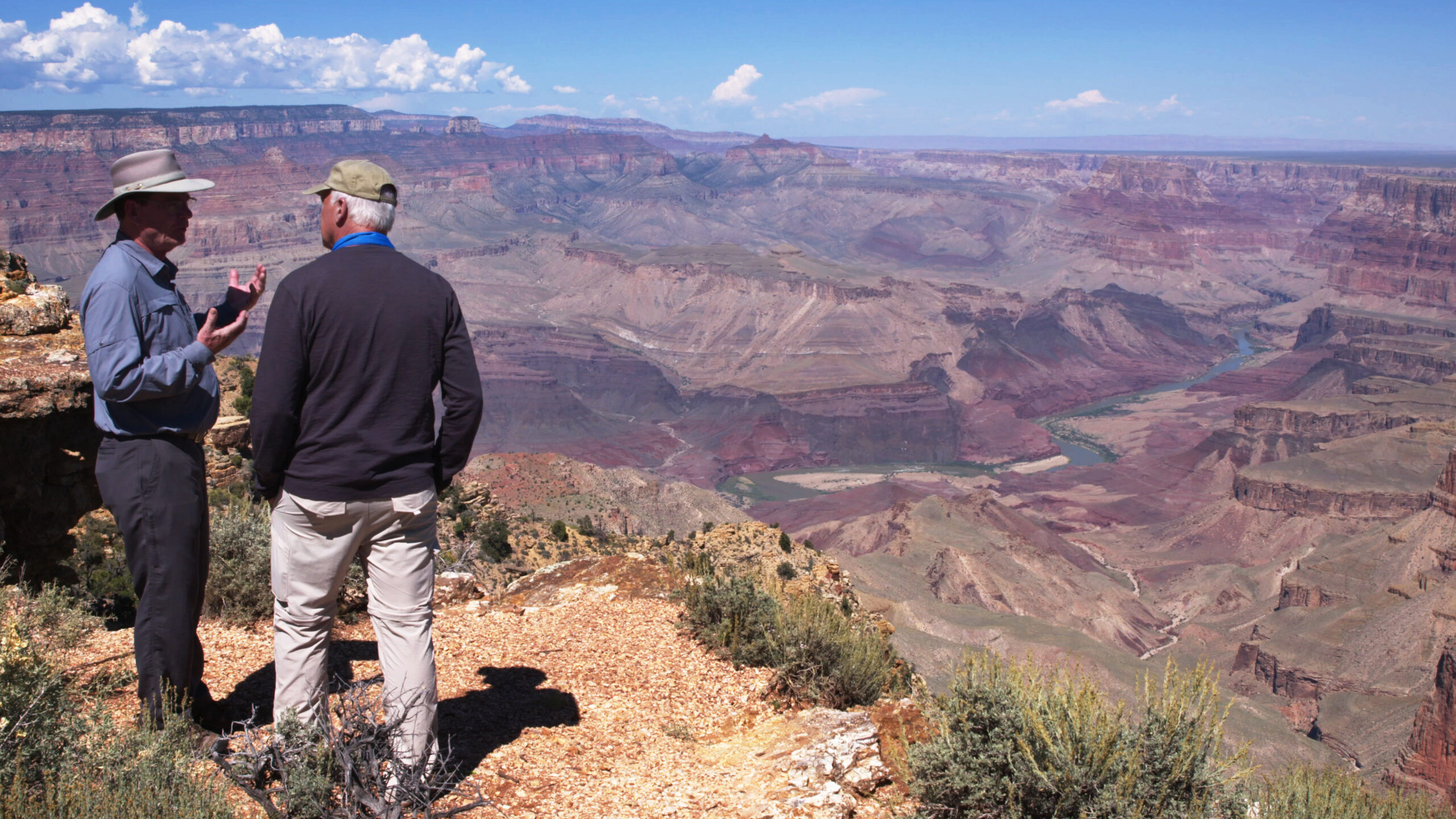 Two men talking at Grand Canyon overlook with expansive rocky landscape and river below.