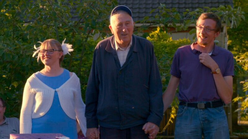 Group of three people standing outside, smiling and holding hands amidst greenery, enjoying a sunny day.