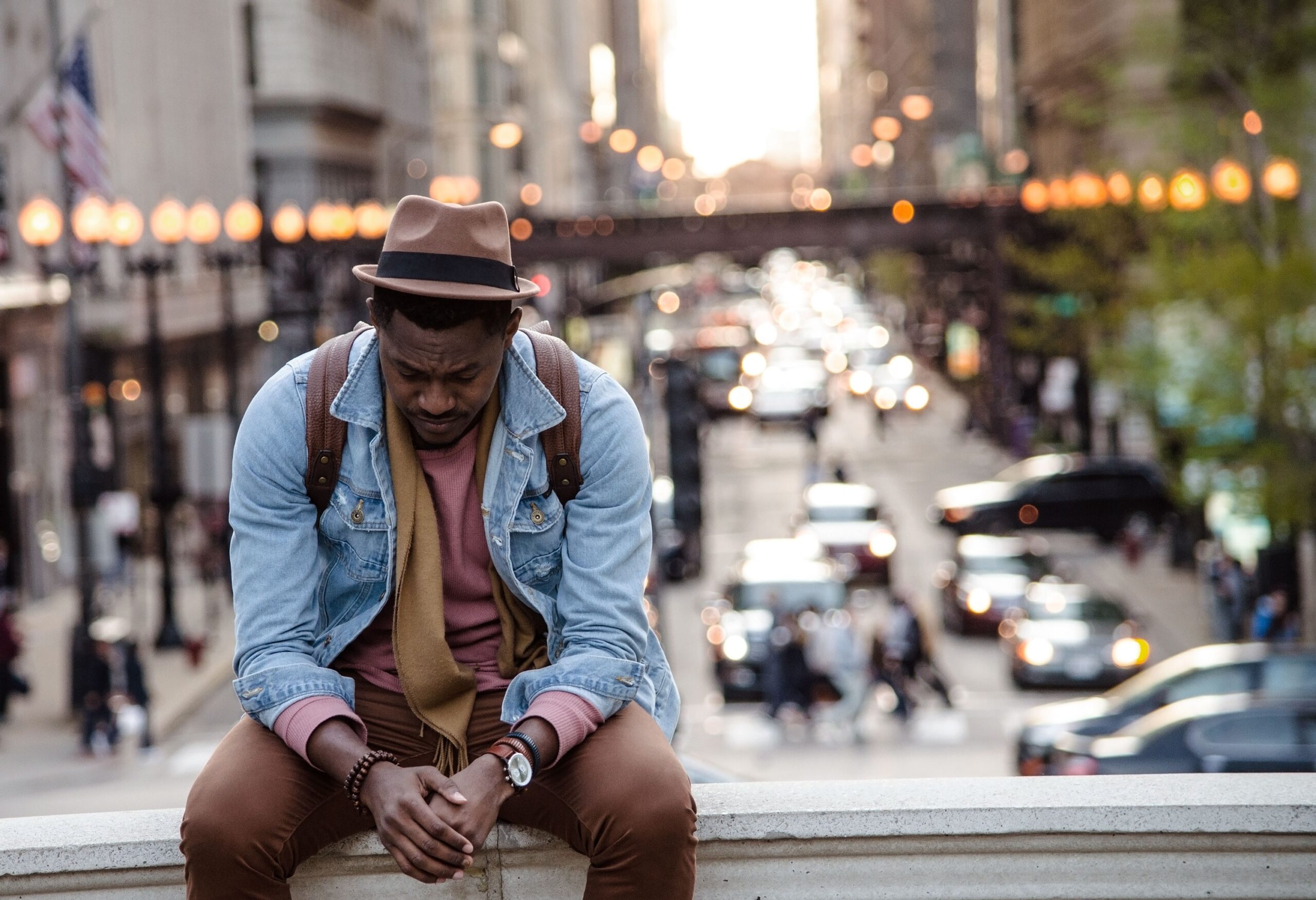 Man sitting pensively on city street ledge, traffic blurred in background, wearing denim jacket and fedora hat.