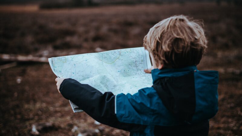 Child exploring nature with a map in hand, wearing a blue jacket in an open field.