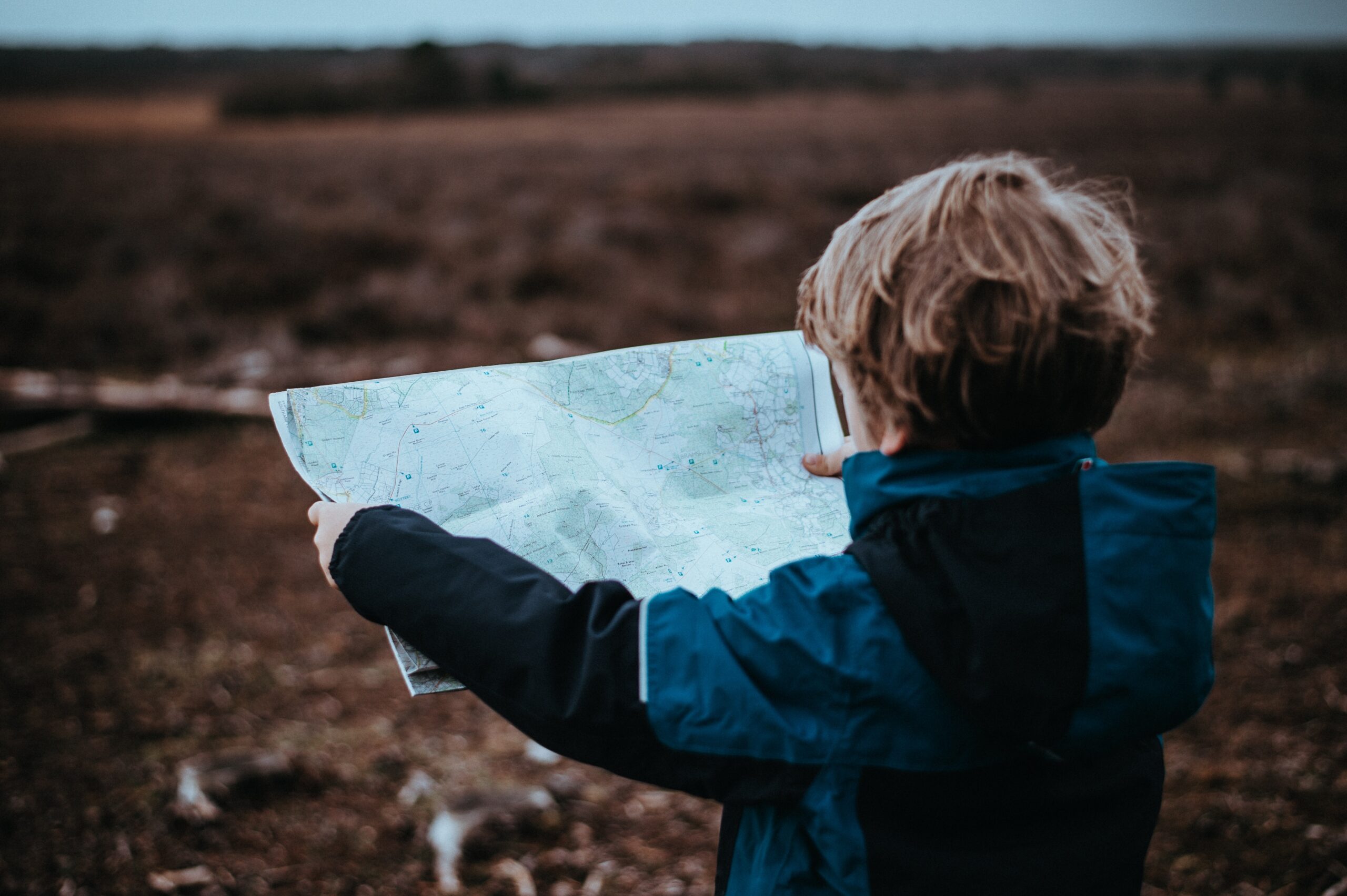 Child exploring nature with a map in hand, wearing a blue jacket in an open field.