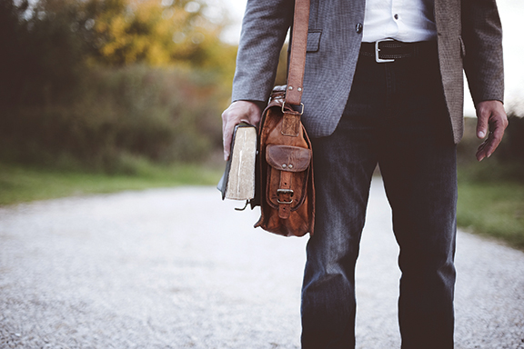 Man holding a leather bag and book, standing on a gravel path in casual attire.