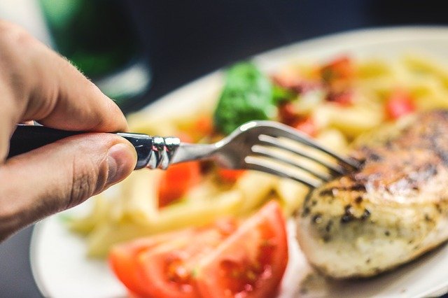 Person eating grilled chicken with pasta and fresh salad on a plate.