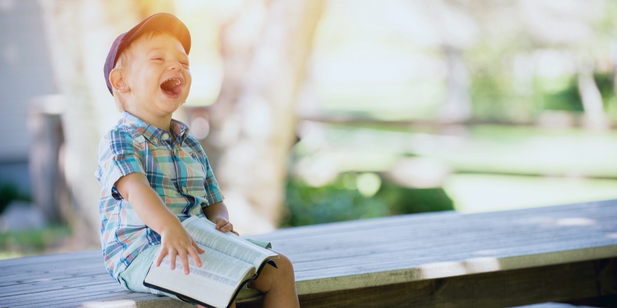A joyful child in a plaid shirt and cap laughs while holding an open book outdoors on a sunny day.