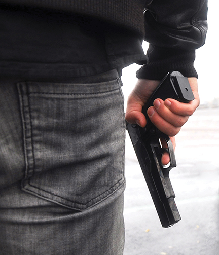 Person holding a handgun behind their back, wearing jeans and a jacket, standing outdoors.