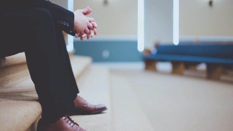 Man in suit sitting on steps with hands clasped, empty church interior in background.