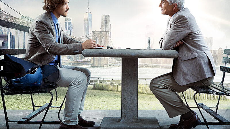Two men conversing at a park table with a cityscape background, sunlight shining through clouds.