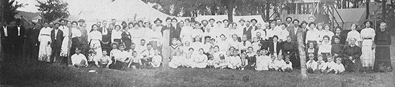 Vintage group photo of people gathered outdoors, early 20th-century attire, trees and tents in background.