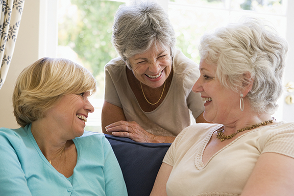 Senior women laughing and chatting together on a couch in a bright, comfortable room.