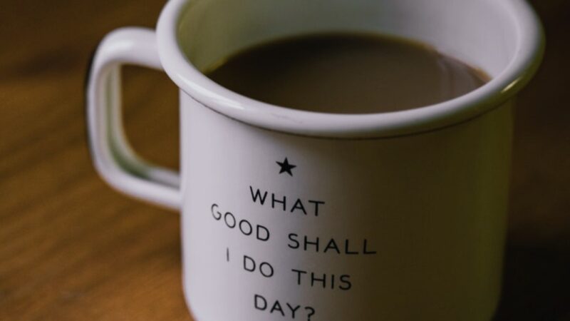 Ceramic mug with motivational quote What good shall I do this day? filled with coffee on wooden table.