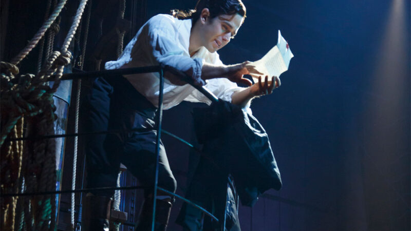 Actor reads a letter dramatically on stage with spotlight, surrounded by a ship's ropes and railings.