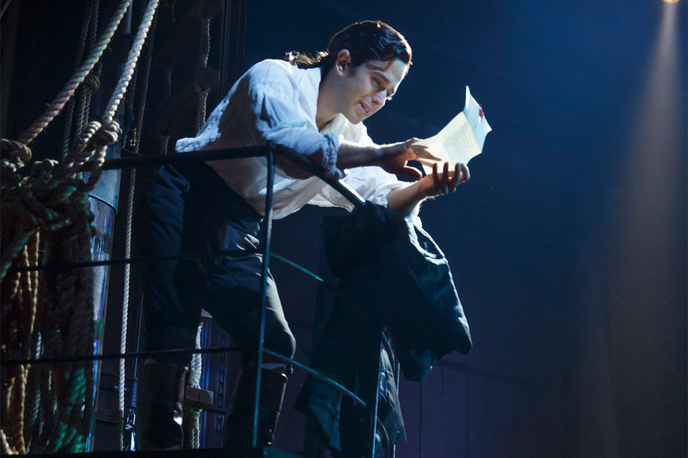 Actor reads a letter dramatically on stage with spotlight, surrounded by a ship's ropes and railings.