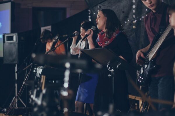 Live band performing on stage with singers and musicians, dimly lit, screen in background showing lyrics.