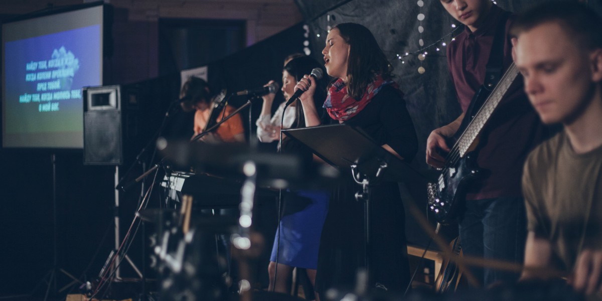 Live band performing on stage with singers and musicians, dimly lit, screen in background showing lyrics.