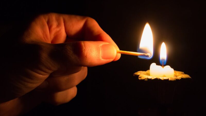 Hand lighting a candle with a matchstick in dark background, creating a warm glow.