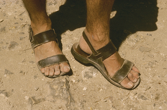 Two feet wearing worn leather sandals standing on a rocky, dusty surface.