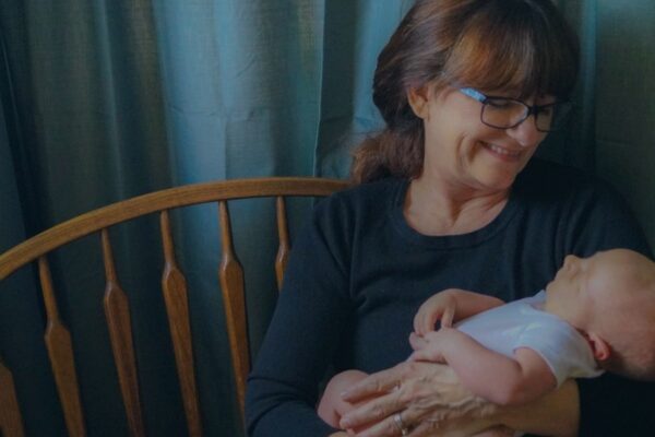 Smiling woman wearing glasses holding a baby in her arms, sitting on a wooden bench with blue curtains in the background.