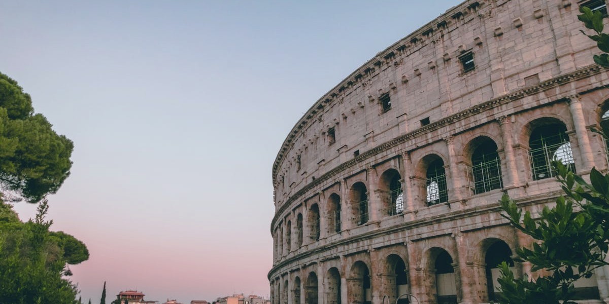Colosseum at sunset with pink sky, Rome's iconic ancient amphitheater, partially obscured by trees.