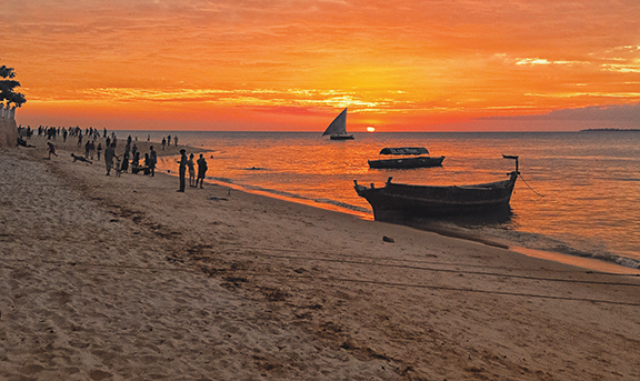 Sunset over a serene beach with boats and people silhouetted against the vibrant orange sky.